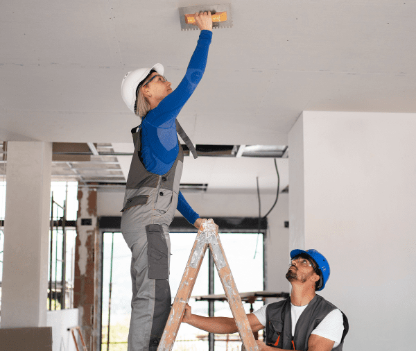 Two construction workers, one standing on a ladder and another assisting, installing or inspecting equipment on a ceiling in a building under renovation. both are wearing hard hats and safety gear.