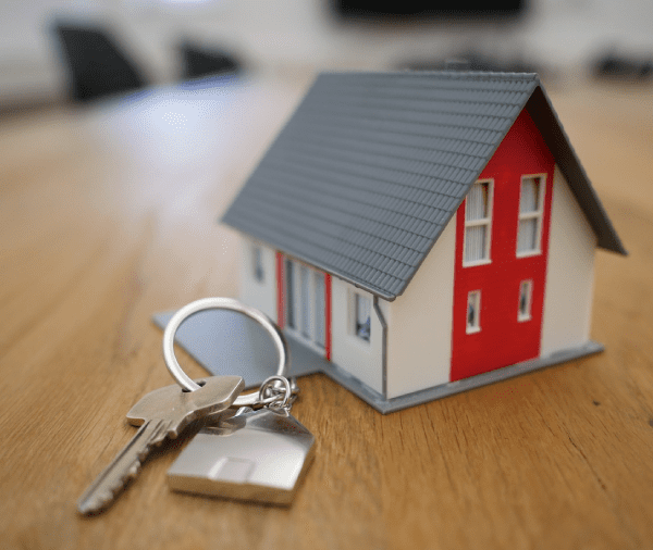 A small model house with red walls and a gray roof, accompanied by keys with a keychain, placed on a wooden table, suggesting themes of real estate or homeownership.