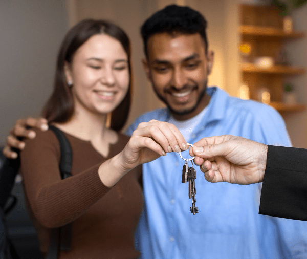 A smiling young couple receives a set of house keys from a person's hand, foreground focused on keys, with a cozy room setting in the background.