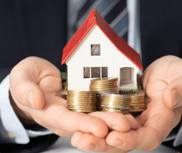 A person in a business suit holds a small model of a house resting on stacked coins, symbolizing real estate investment or homeownership savings.